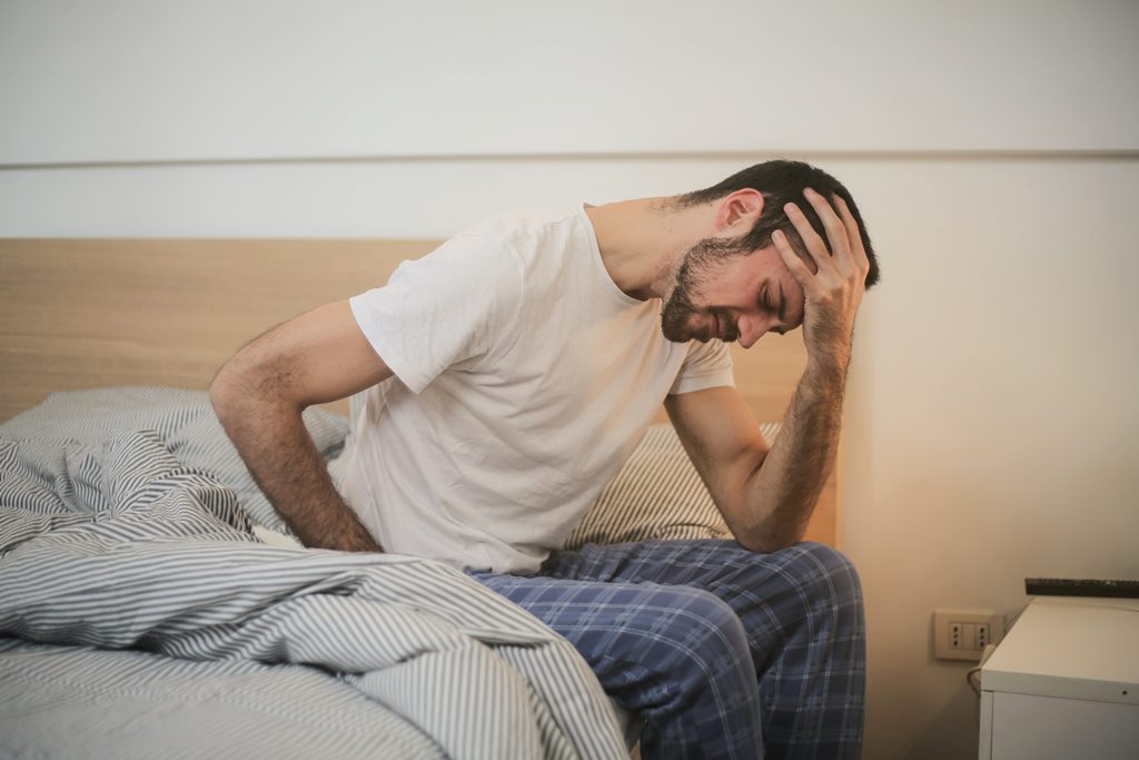 Homem branco de pijama sentado à beira da cama de cabeça baixa, com a mão na testa e expressão de angústia