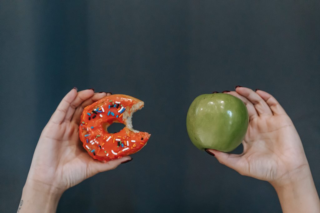 Mãos femininas seguram um donut mordido de um lado e uma maçã verde de outro em alusão a dietas que prometem curar o câncer.